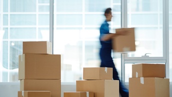  Verschieden große Pakete stehen in einem lichtdurchfluteten Raum mit großen Glasfronten, eine Person in Unschärfe trägt Pakete im Hintergrund