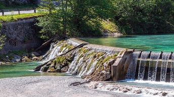Über erhöhte Bebauung in Fluss fließt Wasser, im vorderen Bereich Freies Flussbett, ringsum Bäume
