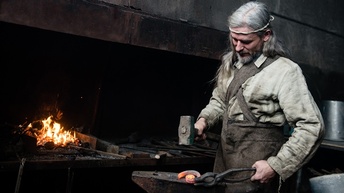 Eine Person steht in einer Schmiede bei einem Amboss und bearbeitet ein glühendes Metallstück mit einem Hammer. Im Hintergrund ist ein Ofen zu sehen, in dem ein Feuer brennt. Die Person hat lange graue Haare und trägt traditionelle Arbeitskleidung.