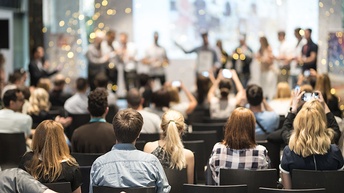 Mehrere sitzende Personen in Rückenansicht, im Hintergrund verschwommen Personen auf Bühne