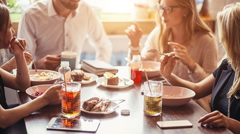 Personen an Tisch sitzend beim Essen. Im Fokus ist ein Holztisch, die Personen sind von der Seite und nur verschwommen zu sehen. Am Tisch sind Stücke von Kuchen und Gläser mit Limonaden platziert.