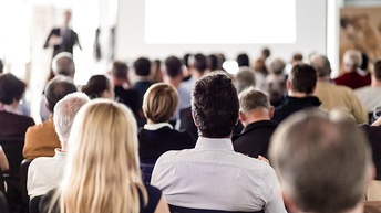 Mehrere sitzende Personen in Rückenansicht, im Hintergrund verschwommen Person auf Bühne