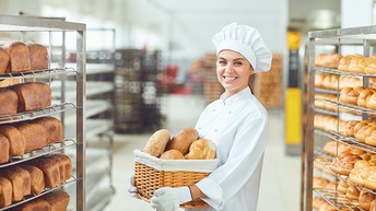 Lächelnde Person in weißem Kochgewand mit Haube hält Korb mit Brotlaiben in Händen