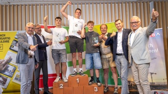 Die drei Gewinner stehen mit erhobenen Händen am Podest und lächeln stolz in die Kamera