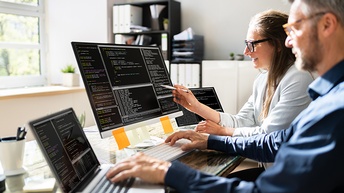 Zwei Personen vor Laptops und Computerbildschirm sitzend und Programmiercodes betrachtend