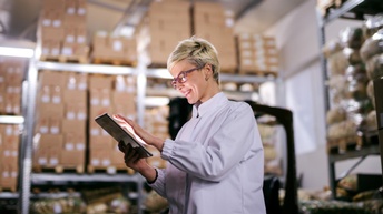  Lächelnde Person mit Brille tippt auf Tablet, im Hintergrund verschwommen Regale mit gestapelten Kartons