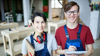 Zwei lächelnde Personen in blauen Arbeitslatzhosen mit Stiften und Notizblöcken in Tischlerwerkstatt stehend