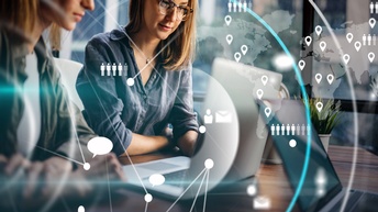 Eine Person sitzt an einem Laptop und arbeitet konzentriert. Über dem Bild liegen transparente Grafiken wie Weltkarte, Standortpunkte und Kommunikationssymbole, die digitale Vernetzung und globale Zusammenarbeit darstellen.