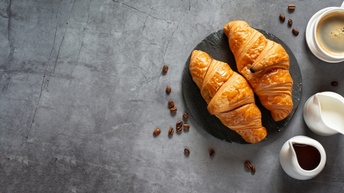 Top-Shot von zwei Croissants auf einem grauen Teller, losen, verstreuten Kaffeebohnen, einer Kanne mit einer dunklen Flüssigkeit, einer Kanne mit einer hellen Flüssigkeit, einer Tasse mit Kaffee