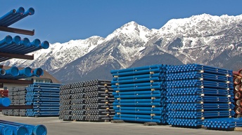 Lagerplatz mit gestapelten blauen Rohren, im Hintergrund beschneite Bergkette und blauer Himmel