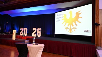 Eine leere Bühne in einem großen Veranstaltungssaal ist zu sehen. Auf der Bühne stehen beleuchtete Zahlen „20 26“. Rechts befindet sich eine große Leinwand mit einem goldenen Adler-Symbol und der Aufschrift „Neujahrsempfang 2026“. Unten rechts ist ein Logo der Wirtschaftskammer Tirol zu sehen. Links steht ein Rednerpult mit Logo. Im Vordergrund stehen zwei Stehtische mit Wasserflaschen und Gläsern. Der Raum ist mit rotem Bühnenvorhang, dunklen Vorhängen und blauer Beleuchtung gestaltet.
