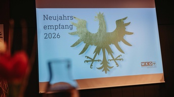 Eine große Projektionsfläche zeigt den Schriftzug „Neujahrsempfang 2026“. Daneben ist ein stilisierter goldener Adler auf hellem Hintergrund zu sehen. Unten rechts befindet sich das Logo der Wirtschaftskammer Tirol. Im Vordergrund ist unscharf ein Glas zu erkennen, was auf eine Veranstaltung mit Publikum hinweist. Der Raum ist warm beleuchtet und wirkt wie ein Veranstaltungssaal.