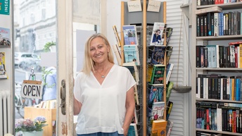 Porträt einer lächelnden Person mit offenen, hellen Haaren, die in einem Geschäft in der geöffneten Tür steht. Hinter der Person sind Regale mit Büchern
