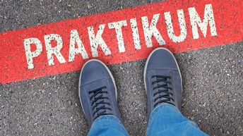 Top-Shot der Turnschuhe einer Person, die auf einer asphaltierten Straße steht. Auf der Straße ist ein rotes Rechteck eingezeichnet. In dem Rechteck steht in weiß der Schriftzug Praktikum