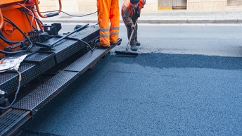Auf einer frisch asphaltierten Straße steht ein großes Fahrzeug zur Asphaltierung. Auf dem Fahrzeug steht eine Person in oranger Hose. Direkt daneben steht eine Person mit dicken Schuhen und Handschuhen, die einen Besen hält