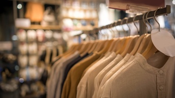 Im Fokus ist ein Kleiderständer, auf dem verschiedene Oberteile in Erdtönen hängen. Der Kleiderständer steht in einem Geschäft