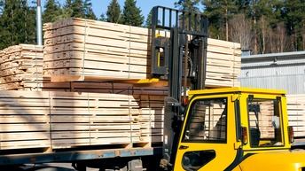 Auf einer erhöhten, blauen Metallplatte stehen viele Stapel heller Holzbretter. Vor der Platte steht ein gelber Gabelstapler, der einen Stapel Holzbretter hochhebt. Im Hintergrund sind hohe, grüne Bäume