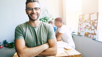Porträt von einer lächelnden Person mit Brille, kurzen Haaren und T-Shirt mit verschränkten Armen, im Hintergrund sitzen weitere Personen an einem Tisch sowie steht eine Pinnwand mit Notizen am Fensterbrett