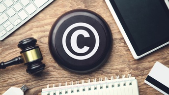 Top-Shot eines runden Objektes in der Mitte, auf dem ein weißes C in einem Kreis ist. Um das Objekt liegen ein Hammer, ein Tablett, eine Computertastatur, ein Notizbuch und ein USB-Stick. Alle Objekte sind auf einer hölzernen Unterfläche