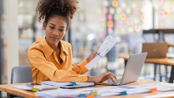 Eine Person mit oranger Bluse und dunklen, geschlossenen Haaren sitzt an einem Schreibtisch. In der linken Hand hält sie Zettel mit Diagramme und Zahlen. Vor ihr liegen viele weitere Papierstapel, auf die sie blickt