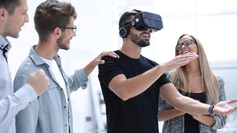Eine Person trägt eine Virtual Reality Brille am Kopf und greift mit den Armen während andere Personen daneben stehen und freudig zusehen
