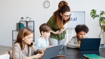 Drei Kinder sitzen an einem Tisch. Jedes Kind hat einen Laptop vor sich. Eine erwachsene Person beugt sich zu den Kindern. Sie befinden sich in einem hellen Raum. Im Hintergrund sind ein großer Wandbildschirm, eine Pflanze und ein Regal zu sehen.
