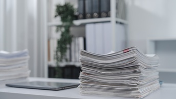 Auf einem weißen Tisch ist ein Dokumentenstapel. Daneben sind ein Tablet und ein weiterer Dokumentenstapel. Im Hintergrund ist ein Regal mit Ordnern