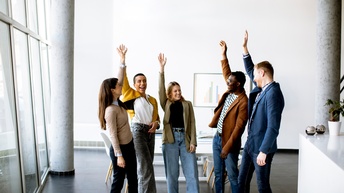Mehrere Personen stehen freudig in einem hellen, modernen Büroräumlichkeit und heben motiviert ihre Hände nach oben