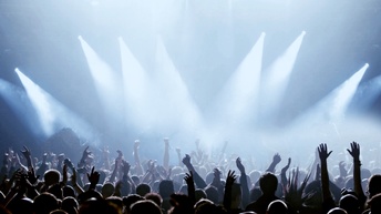 Viele Personen, Fans, stehen vor einer Bühne, die mit Scheinwerfern beleuchtet ist