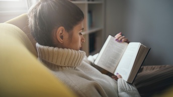 Seitliche Aufnahme einer Person mit geschlossenen Haaren und dickem Pullover, die auf einem gelben Polstermöbelstück sitzt und in einem Buch liest