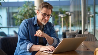 Eine Person mit Brille, Bart und kurzen Haaren sitzt an einem Tisch. Vor ihr ist ein aufgeklappter Laptop, auf dessen Bildschirm sie blickt