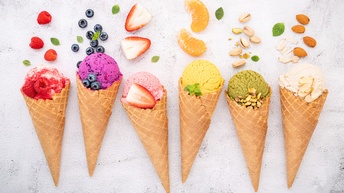 Auf hellem Untergrund liegen sechs Eistüten nebeneinander. In jeder Tüte ist eine Eiskugel mit einer anderen Farbe. Vor jeder Kugel liegt ein Obst oder eine Nuss, von links nach rechts Himbeeren, Blaubeeren, Erdbeeren, Mandarinen, Pistazien, Mandeln