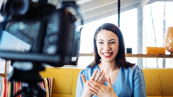 Lächelnde Person mit dunklen Haaren und roten Lippen strahlt in die Kamera, die im Vordergrund unscharf in Bild kragt