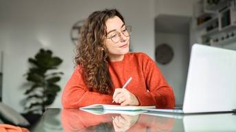 Eine Person mit lockigem, braunem Haar und Brille sitzt an einem Tisch und blickt auf den Bildschirm eines Laptops. Die Person hält einen Stift. Vor ihr liegt ein Notizheft
