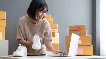 Lächelnde Person steht an Tisch, blickt auf aufgeklapptes Notebook und hält in einer Hand weißen Sneaker, im Hintergrund gestapelte Kartonagen