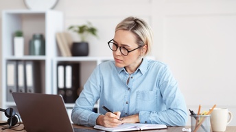 Person mit Brillen sitzt an Tisch, blickt auf aufgeklapptes Notebook vor sich und nimmt Notizen in Buch vor, am Tisch Becher mit Stiften, Tasse und Headphones liegend, im Hintergrund verschwommen Regal mit Aktenordnern