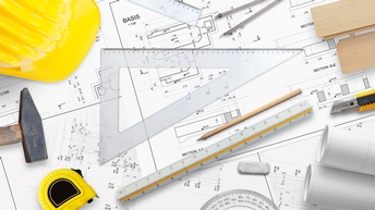 Top-Shot eines weiß-bedruckten Papiers mit verschiedenen Zahlen, Strichen und Linien. Auf dem Papier liegen Lineale, Stifte, ein Hammer, ein Maßband, Papierrollen, ein Zirkel, ein Stanleymesser, ein Helm und Holzbretter.