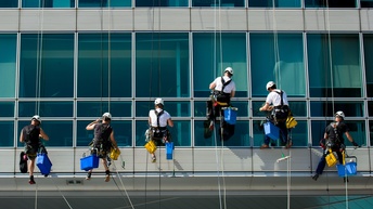Mehrere Personen in Arbeitskleidung und mit Helmen seilen sich an Glasfassade ab und reinigen Fenster