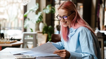 Person mit langen rosaroten Haaren und Brille blickt lächelnd auf Papierdokumente