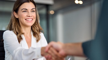 Person mit langen dunklen Haaren sitzt an einem Tisch und hält ein Dokument mit einer Hand während sie freudig die Hand einer Person in der Unschärfe reicht, die gegenüber sitzt