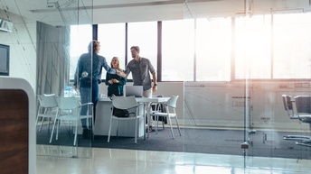 In Meetingraum mit verglasten Wänden schütteln sich zwei Personen über den Tisch die Hand, eine weitere Person steht daneben