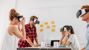 Personen mit Virtual Reality Brillen befinden sich in einem Büroraum mit Tisch auf dem Tassen, Brillen, Unterlagen sowie ein Laptop steht, im Hintergrund befindet sich ein Whiteboard mit Notizen