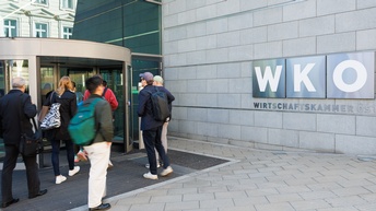 Rückenansicht mehrerer Menschen, die durch die Drehtür eines Gebäudes gehen. Die Mauer des Gebäudes besteht aus grauen, rechteckigen Betonsteinen, auf diesen ist das Logo der Wirtschaftskammern Österreich mit dem Schriftzug WKO auf Metallschilder gedruckt. 