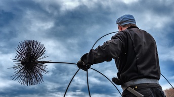 Eine Person in schwarzer Kleidung steht mit dem Rücken zur Kamera und hält in der linken Hand einen Kaminbesen. Im Hintergrund ist der bewölkte, graue Himmel