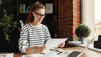 Eine Person mit Brille sitzt an einem Tisch aus hellem Holz vor einem aufgeklappten Laptop. Neben diesem sind verschiedene Dokumente mit Statistiken. Die Person hält einen Zettel, auf den sie blickt. Im Hintergrund ist eine Ziegelwand und ein Regal
