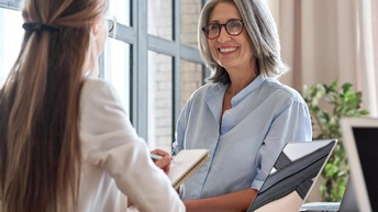 Person mit grauen Haaren und Brillen lächelt Person mit langen Haaren vor aufgeklappten Notebook an