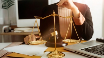 Eine Person sitzt an einem Tisch. Mit der rechten Hand hält sie einen Richterhammer. Vor ihr liegen Unterlagen. Auf dem Tisch steht ein aufgeklappter Laptop und eine goldene Waage