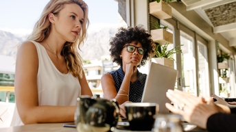 An einem Tisch sitzen sich vier Personen gegenüber. Von den Personen rechts sind nur die Hände im Bild. Die anderen Personen blicken in den rechten Bildbereich. Vor der linken Person steht ein aufgeklappter Laptop.