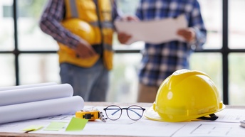 Im Vordergrund liegt ein Bauplan auf einem Tisch. Auf dem Plan liegen Papierrollen, eine Brille, ein Bauhelm und ein Maßband. Im Hintergrund stehen zwei Personen nebeneinander. Die linke hält einen Plan