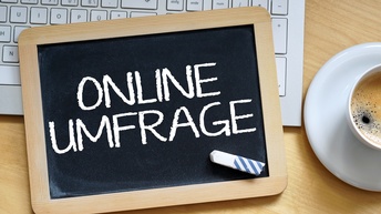Auf einer Schiefertafel steht in Versalien das Wort Onlineumfrage. Darauf liegt ein Stück Kreide. Unter der Schiefertafel ist die Tastatur eines Laptops. Daneben in rechter Bildhälfte ist eine Untertasse und eine Tasse mit Kaffee